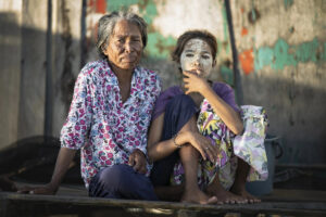 Malaysia, Boheyan island, 2024-05-10. Young girl and old Bajau woman sat together in front of their stilt house. Photogr