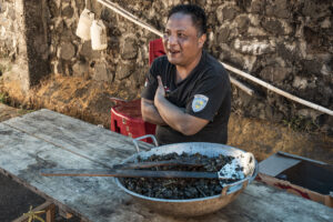 Indonesia, Tomohon, 2026-09-13. Ipam Daulima sits outside his home behind a wok of RW, dog meat cooked with Minahasa spi