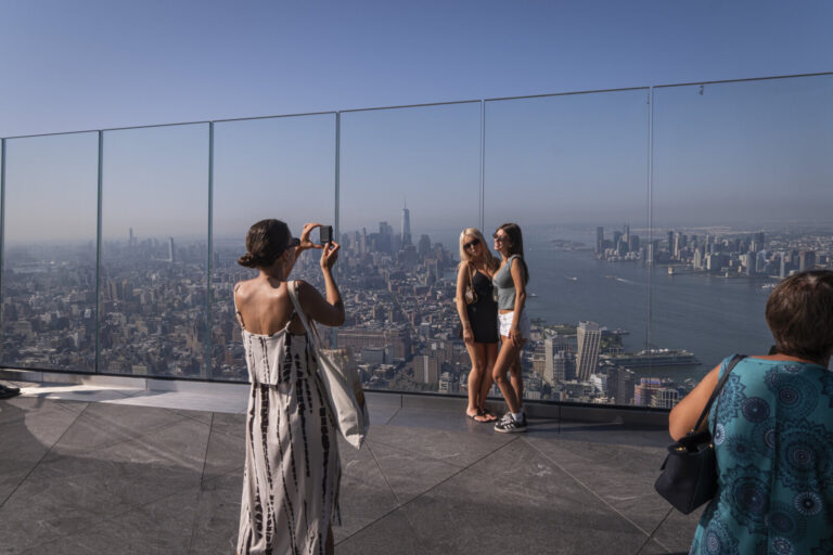 United States, New York, 2026-08-07. A woman photographs two friends against the skyline of Lower Manhattan. Photograph 