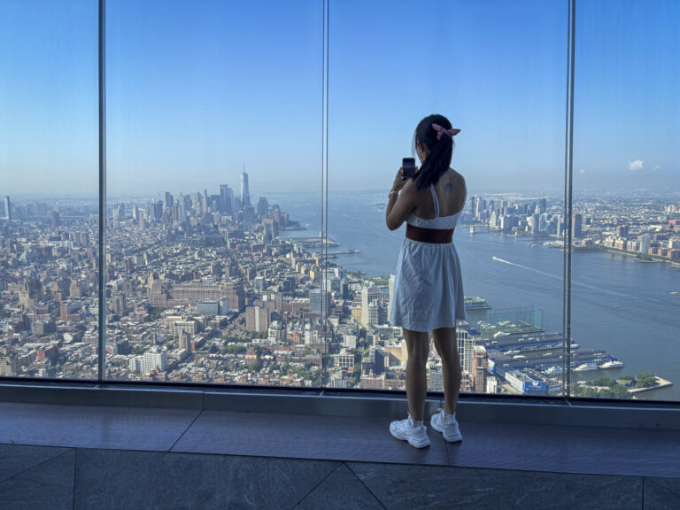 United States, New York, 2026-08-07. A visitor photographs Lower Manhattan through the glass wall of the observation dec