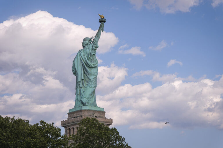 United States, New York, 2026-08-15. The Statue of Liberty seen from behind, under a sky dotted with clouds. Photograph 