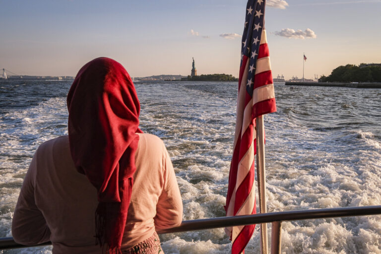 United States, New York, 2026-08-15. A veiled passenger and the American flag on the deck of the ferry, facing the statu