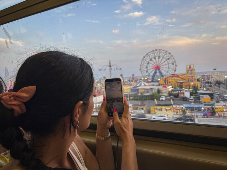 United States, New York, 2026-08-08. A passenger photographs the amusement park from a train window at dusk. Photograph 