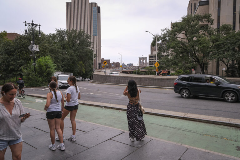 United States, New York, 2026-08-16. Passers-by wait near the approach of the Brooklyn Bridge as cars go by. Photograph 