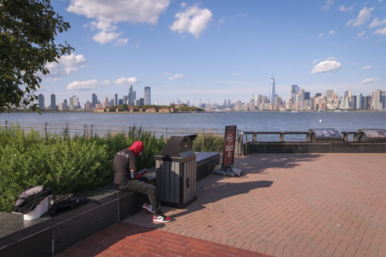 United States, New York, 2026-08-15. A man sits facing the skyline of Manhattan, on the promenade of Liberty Island. Pho
