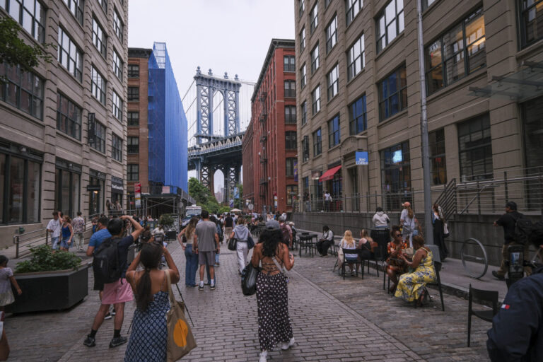 United States, New York, 2026-08-16. Visitors sit at terraces on Washington Street in Dumbo under the Manhattan Bridge. 
