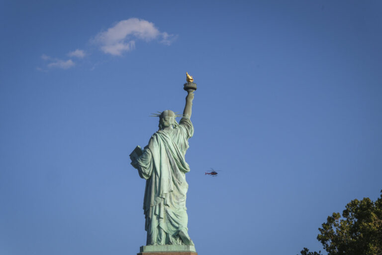 United States, New York, 2026-08-15. The Statue of Liberty seen from behind, a helicopter crossing the blue sky. Photogr