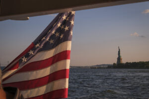 United States, New York, 2026-08-15. The American flag of the ferry and, in the distance, the Statue of Liberty at sunse