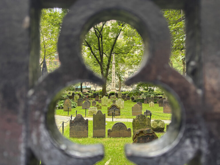 United States, New York, 2026-08-06. The Trinity Church graveyard seen through an opening in its iron fence. Photograph 