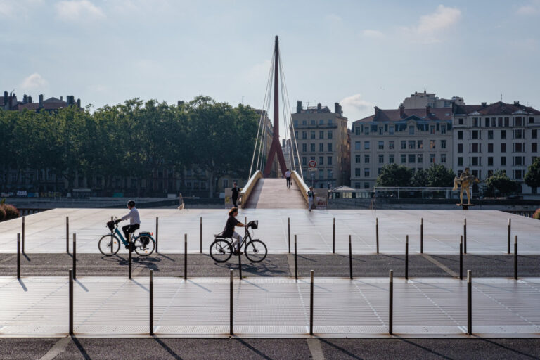 France, Lyon, 2024-06-26. Two cyclists passing on the courthouse square with the footbridge in the background. Photograp