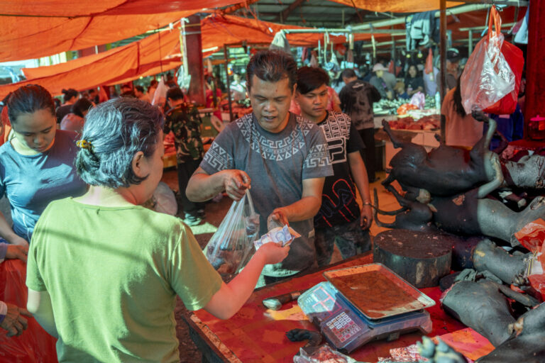 Indonesia, Tomohon, 2026-09-12. Vendor handing a bag of dog meat to a customer paying in cash, dog carcass hanging in th