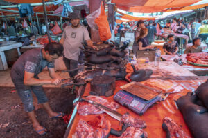 Indonesia, Tomohon, 2026-09-12. Butcher cutting a dog carcass with a knife on a chopping block, second vendor nearby, cr