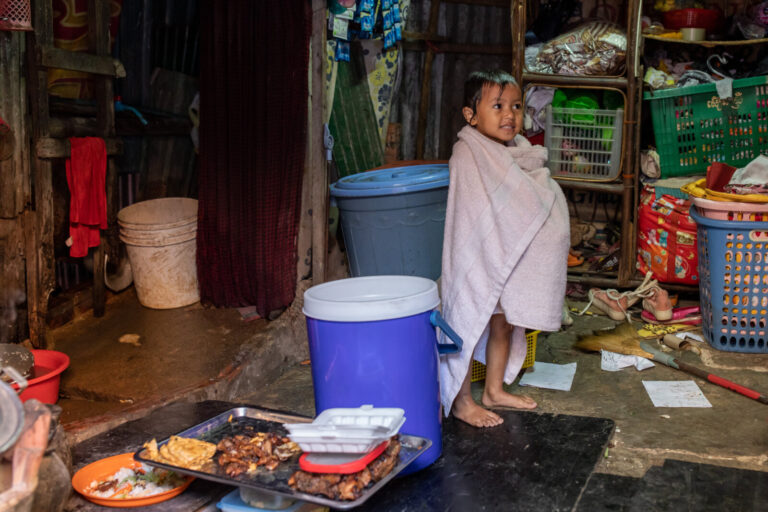 Cambodia, Phom Penh, Tuol Kok, 2019-11-29. A child in a bath towel, inside a dwelling amidst cans, baskets and trays of 