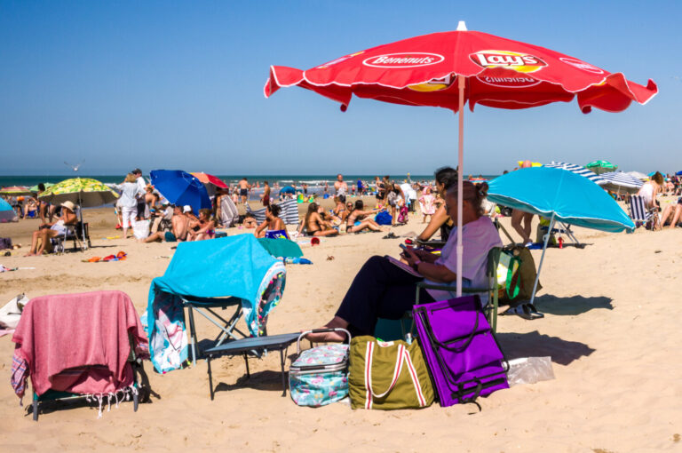 France, Bray-Dunes, 2026-08-12. Deck chairs and bags sit under a red parasol on a crowded summer beach. Visitors bring p