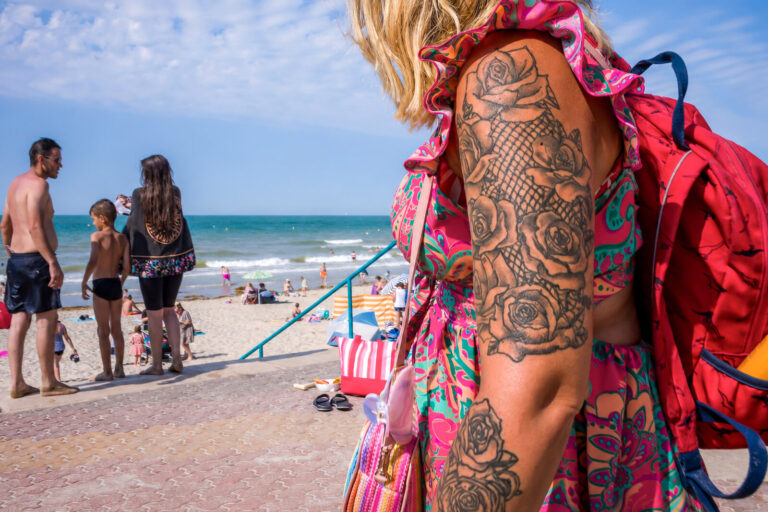 France, Bray-Dunes, 2026-08-15. A tattooed person with a red backpack stands in the foreground as beachgoers walk near t