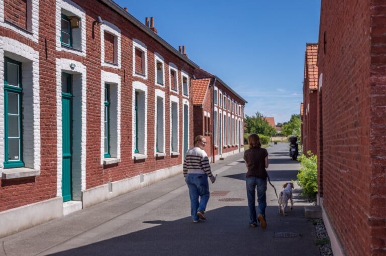 France, Bruay-la-Buissiere, 2026-06-29. Two people walk a dog along a lane bordered by restored brick miners houses. The