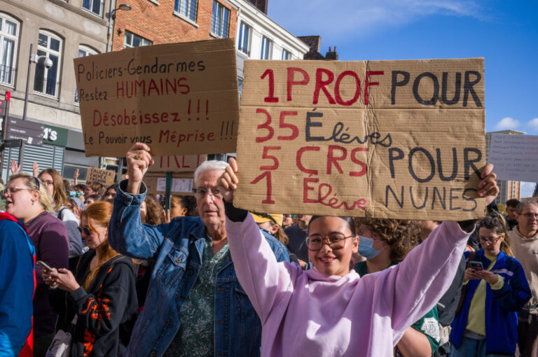 France, Lille, 2026-10-17. Two protesters hold cardboard signs comparing teachers and riot police and calling on police 