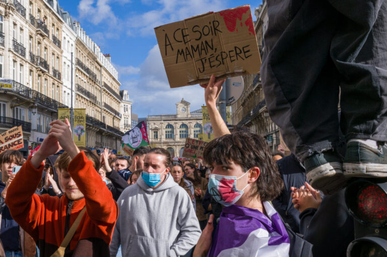 France, Lille, 2026-10-17. A masked protester raises a cardboard sign addressing a mother, while another participant cla